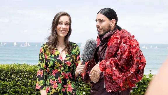 Alina Stiegler steht mit einem Mikro neben Conan Osíris aus Portugal. Im Hintergrund das Meer.