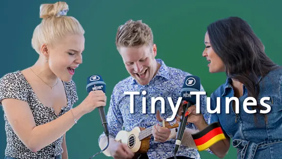 Eine blonde und eine dunkelhaarige Frau singen jeweils in ein Mikrofon, in der Mitte spielt ein Mann auf einer Ukulele.